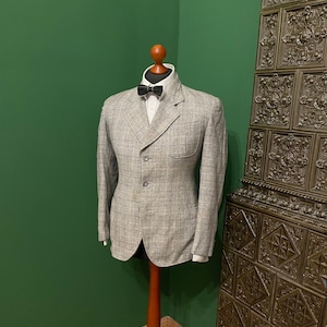 Könnte beinhalten: Ein grauer karierter Blazer mit einem weißen Hemd mit Kragen und einer schwarzen Fliege. Der Blazer befindet sich auf einem hölzernen Mannequin vor einer grünen Wand.
