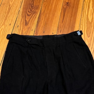 Op de afbeelding: Close-up van een zwarte broek met plooien en een knoopsluiting. De broek heeft een riemlus met een blauw accent. De stof lijkt een donkere, effen kleur te hebben. De broek ligt op een houten oppervlak.