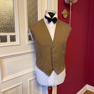 Könnte beinhalten: Eine braun-gold gestreifte Weste mit schwarzer Fliege und weißem Hemd mit Kragen. Die Weste hat eine Knopfleiste und wird auf einer Schaufensterpuppe präsentiert. Das Outfit befindet sich vor einer weißen Tür und einer roten Wand.