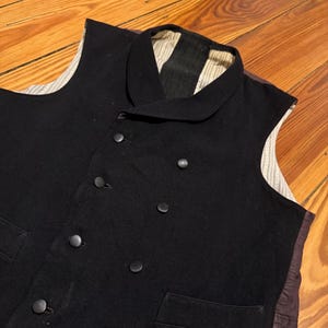 May include: A black vest with a notched collar and six dark buttons. The vest has a pocket on the lower left side and a striped lining visible at the collar and armholes. The vest is made of a dark, possibly wool, material.