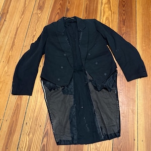 May include: A black formal jacket with a double-breasted front and long tails. The jacket is made of a dark fabric and is lying on a wooden floor. The jacket has a classic design.