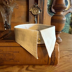 Puede incluir: Un cuello blanco desmontable con una parte delantera puntiaguda, asegurado por un botón dorado. El cuello está hecho de una tela texturizada y descansa sobre un libro antiguo. El fondo incluye una mesa de madera y decoración antigua.