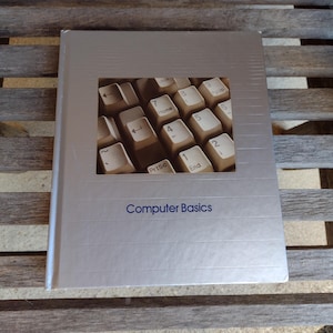 May include: A silver book titled "Computer Basics" with a rectangular cutout showing a close-up of computer keyboard keys. The book is on a wooden bench.