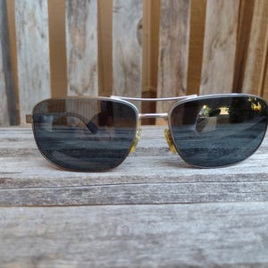 Może przedstawiać: Okulary przeciwsłoneczne w stylu Aviator ze srebrną oprawką i ciemnymi soczewkami. Okulary spoczywają na zwietrzałej drewnianej powierzchni, odbijając otoczenie. Soczewki mają lekkie przejście tonalne, a design jest klasyczny.