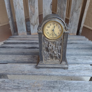 Könnte beinhalten: Eine silberfarbene Uhr mit goldenem Zifferblatt und schwarzen Zeigern. Die Uhr hat die Form eines kleinen Turms mit einem dekorativen Design auf der Vorderseite. Die Uhr steht auf einer Holzoberfläche.