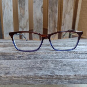 以下が含まれることがあります： 茶色と青色の縁取りの長方形フレームの眼鏡。 フレームの上部は茶色で、下部で青色に変わります。 眼鏡は風化した木製の表面に置かれています。