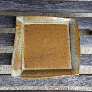 以下が含まれることがあります： 薄い茶色の釉薬がかかった正方形のセラミック製のサービングディッシュ。縁がわずかに盛り上がっており、斑点模様で、微妙な凹凸があります。