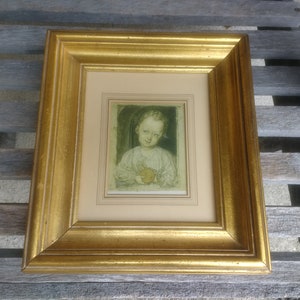 May include: A gold-framed picture with a white mat and a sepia-toned image of a young person holding an apple. The image is set against a green background.