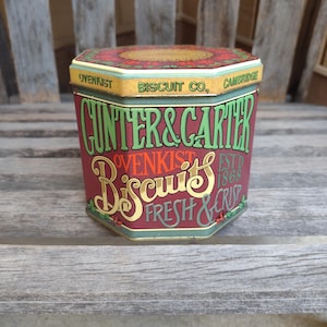 Puede incluir: Una lata de galletas vintage, hexagonal, con fondo rojo y letras doradas. La lata dice "GUNTER & CARTER OVENKIST BISCUITS FRESH & CRISP" y "ESTD 1868". La tapa está decorada con un diseño floral.