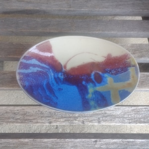 Puede incluir: Un plato ovalado de cerámica blanca con un diseño abstracto azul, amarillo y rojo. El plato está esmaltado y tiene un acabado liso.