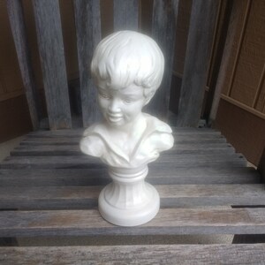 May include: White ceramic bust sculpture of a young boy with short hair. The bust is on a pedestal with a fluted base.