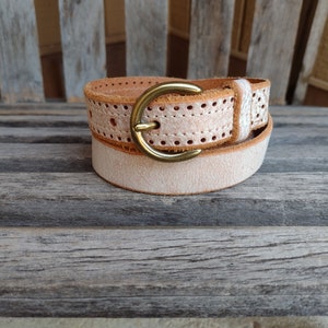 May include: A light brown leather belt with a brass buckle and a perforated design. The belt is rolled up and sitting on a wooden surface.