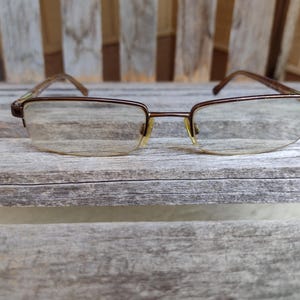 Może przedstawiać: Okulary z brązową metalową oprawką i przezroczystymi soczewkami. Okulary spoczywają na zwietrzałej drewnianej powierzchni. Soczewki mają lekką żółtą poświatę.