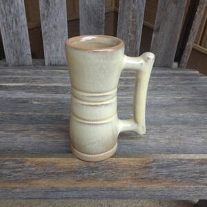 May include: A light yellow ceramic mug with a wide handle. The mug has a textured surface and is decorated with two brown bands.