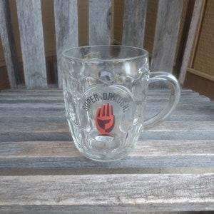 Puede incluir: Una jarra de cerveza de vidrio transparente con asa. La jarra tiene un diseño en relieve y dice "Super Draught" con una mano roja sosteniendo un vaso de cerveza.