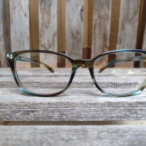 May include: A pair of eyeglasses with a brown and teal patterned frame. The lenses are clear, and the brand name "TIFFANY & CO." is visible on the right lens. The glasses rest on a weathered wooden surface.