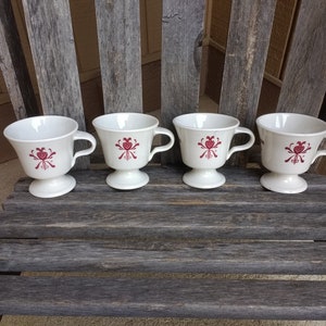 Op de afbeelding: Vier witte keramische theekopjes met rode bloemenmotieven. De kopjes hebben een voet en een handvat. De kopjes zijn in een rij op een houten oppervlak gerangschikt.