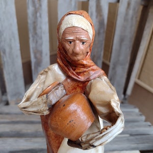 Puede incluir: Una figurita hecha a mano de una mujer mayor que lleva un chal marrón y blanco, sosteniendo una olla de barro marrón. La figurita está hecha de papel maché y tiene un aspecto rústico y vintage.