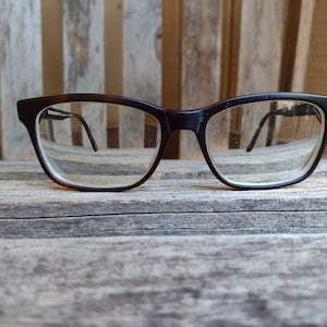 May include: A pair of black framed spectacles with clear lenses. The glasses are resting on a wooden surface.