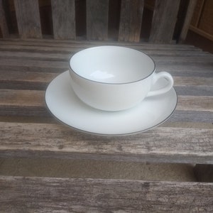 Könnte beinhalten: Eine weiße Teetasse und Untertasse mit einem dünnen schwarzen Rand. Tasse und Untertasse stehen auf einer Holzoberfläche.