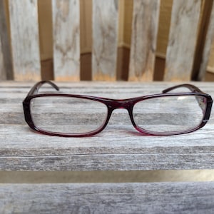 Könnte beinhalten: Eine rechteckige Brille mit einem dunkelburgunderfarbenen Rahmen liegt auf einer verwitterten Holzoberfläche. Die Gläser sind klar, und die Bügel sind leicht gebogen. Die Brille dient wahrscheinlich zur Sehkorrektur.