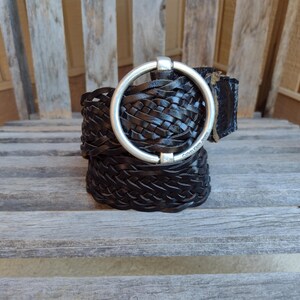 May include: Black woven leather belt with a large silver-tone circular buckle. The belt features a braided design and the words "Calvin Klein Jeans" are visible on the buckle. The belt is displayed on a wooden chair.