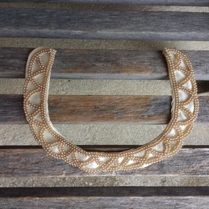 May include: A white fabric choker necklace with a beaded design. The beads are a light gold color and are arranged in a geometric pattern.