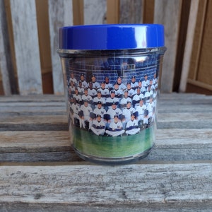 May include: A clear glass jar with a blue lid, containing a photo of a baseball team. The photo shows a group of men wearing white and blue uniforms, sitting on a green field.