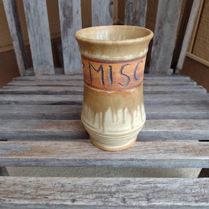 Pode incluir: Um pote de cerâmica com um esmalte marrom e branco. O pote tem a palavra "MISC" escrita em letras marrons.