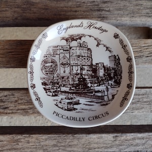 May include: A white ceramic plate with a brown design depicting a street scene in London, England. The plate features the text "England's Heritage" and "Piccadilly Circus".