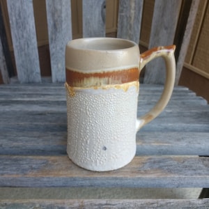 May include: A ceramic beer stein with a brown and white glaze. The stein has a textured surface and a handle.