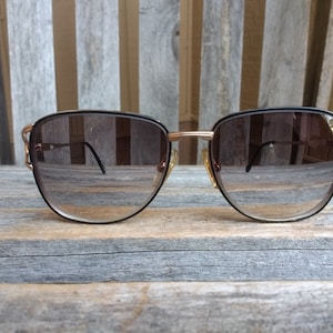 May include: Vintage sunglasses with a black frame and gold-tone accents. The lenses are a gradient brown color. The sunglasses have a unique design on the temples and are set against a weathered wooden background.