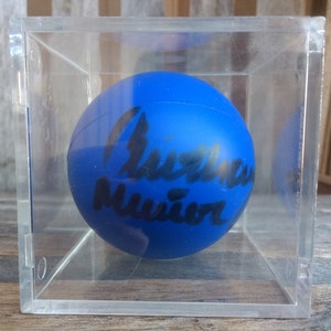 May include: A blue bouncy ball with the signature "Christine Muñiz" written in black ink. The ball is encased in a clear plastic cube.