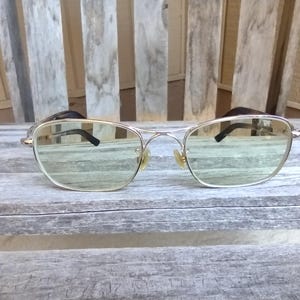 May include: A pair of aviator-style sunglasses with a silver metal frame and light green lenses. The sunglasses are resting on a weathered wooden surface, with a wooden background.
