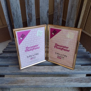 May include: Two gold-coloured picture frames with a pink and white patterned design. The frames are labelled "Decorator Photoframe" and have the dimensions "5 in x 7 in".