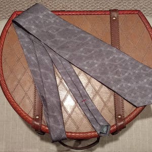 May include: A gray patterned silk tie on a brown leather background with a red and brown trim.