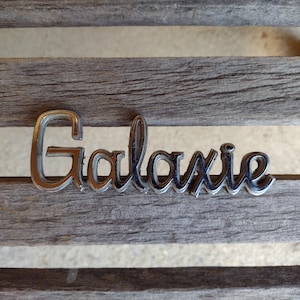 May include: A chrome "Galaxie" emblem, with a cursive font, rests on a weathered wooden surface. The emblem's metallic finish contrasts with the gray wood grain, creating a vintage aesthetic. The word "Galaxie" is clearly visible.