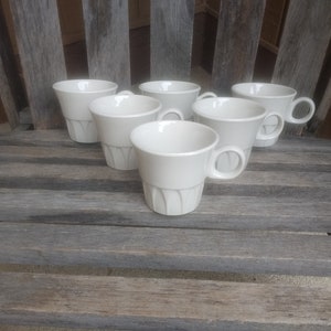 Könnte beinhalten: Sechs weiße Keramik-Kaffeetassen mit geriffeltem Design. Die Tassen sind versetzt auf einer Holzoberfläche angeordnet.