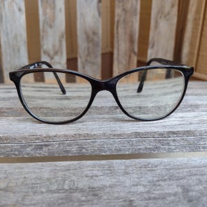 Może przedstawiać: Okulary w czarnej oprawie o klasycznym kształcie kociego oka. Soczewki są przezroczyste, a okulary leżą na tle zwietrzałego drewna. Błyszczące czarne oprawki zapewniają prosty i elegancki wygląd.