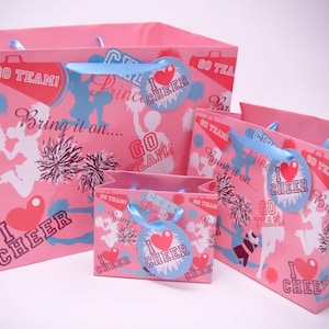 Può includere: Quattro sacchetti regalo rosa con silhouette di cheerleader bianche, pompon e il testo "Go Team!" e "I Love Cheer". I sacchetti hanno manici in nastro blu e etichette.