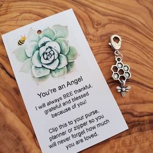 Puede incluir: Una tarjeta blanca con una ilustración de acuarela de una planta suculenta y una abeja. La tarjeta tiene el texto "You're an Angel" y un mensaje sobre estar agradecido y agradecido. Un amuleto plateado con un panal y una abeja está unido a la tarjeta.