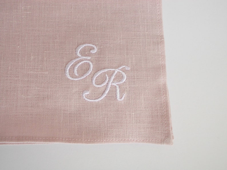 May include: A pale pink linen napkin with white embroidered initials "E & R" in a cursive font.