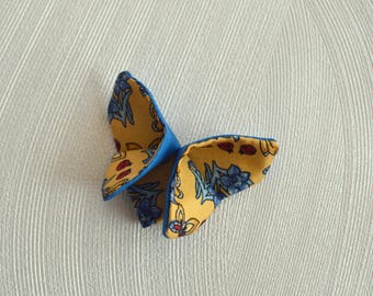 May include: A handmade butterfly-shaped fabric ornament. The wings are a vibrant yellow with a floral pattern in blue and red, outlined with a bright blue edge. The ornament is set against a neutral background.