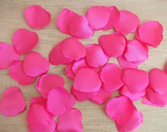 Puede incluir: Un montón grande de pétalos de rosa de satén rosa brillante. Los pétalos están esparcidos sobre una superficie de madera marrón claro.