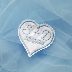 May include: White heart-shaped fabric patch with blue embroidery. The patch features the initials "S + D" and the date "10/15/24".