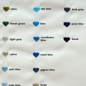 May include: A fabric swatch featuring embroidered heart shapes in various colors, including emerald green, steel blue, and dark oak. Each heart is labeled with its corresponding color name, such as "sky blue" and "navy blue."