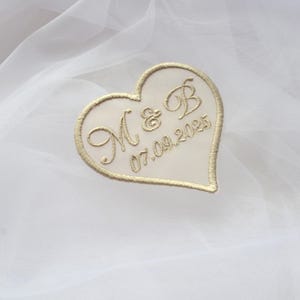 以下が含まれることがあります： 金色の刺繍で「M & B」のイニシャルと日付「07.09.2025」が施された、クリーム色のハート型のパッチ。 パッチは金色の縁取りがあり、白のテクスチャード加工された背景に配置されています。