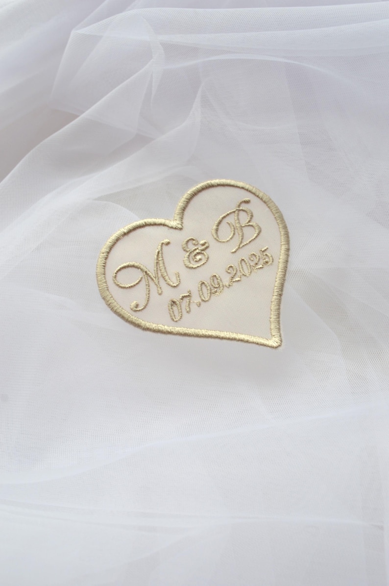May include: A heart-shaped patch with gold embroidered initials "M & B" and the date "07.09.2025". The patch is cream-colored with a gold border, set against a white, textured background.