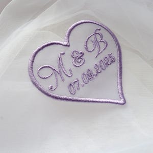 Puede incluir: Un parche bordado en forma de corazón con las iniciales "M & B" y la fecha "07.08.2025" en hilo lavanda. El parche está sobre un fondo de tela de tul blanca. El parche tiene un borde lavanda.