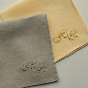 May include: Two square fabric napkins, one in a pale yellow and one in a muted grey, each featuring a delicate cursive monogram embroidery. The yellow napkin has a golden yellow thread monogram, while the grey napkin has a matching grey thread.